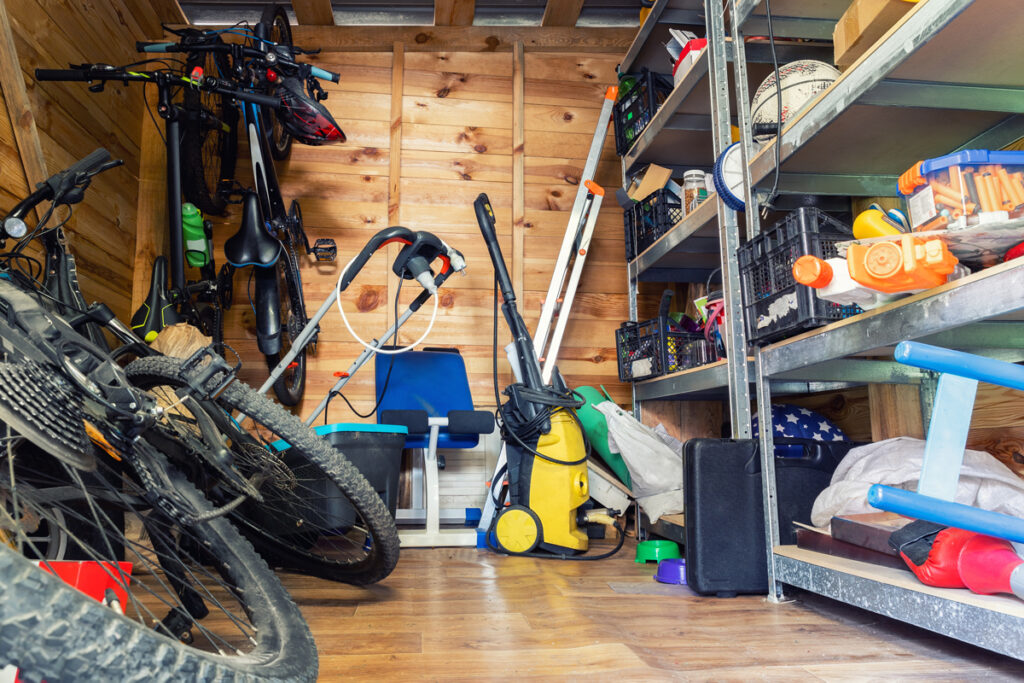 A cluttered wooden shed interior with bicycles, a lawn mower, garden tools, a blue chair, and shelves filled with boxes, toys, and various household items.