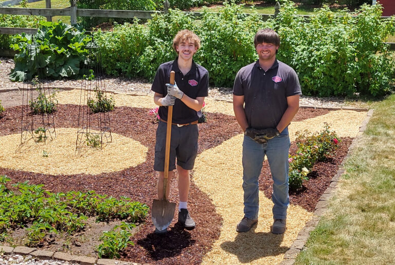 Two young men stand in a well-maintained garden, one holding a shovel and wearing gloves. Both are smiling, dressed in matching dark shirts with logos, surrounded by green plants and a mulch pathway.