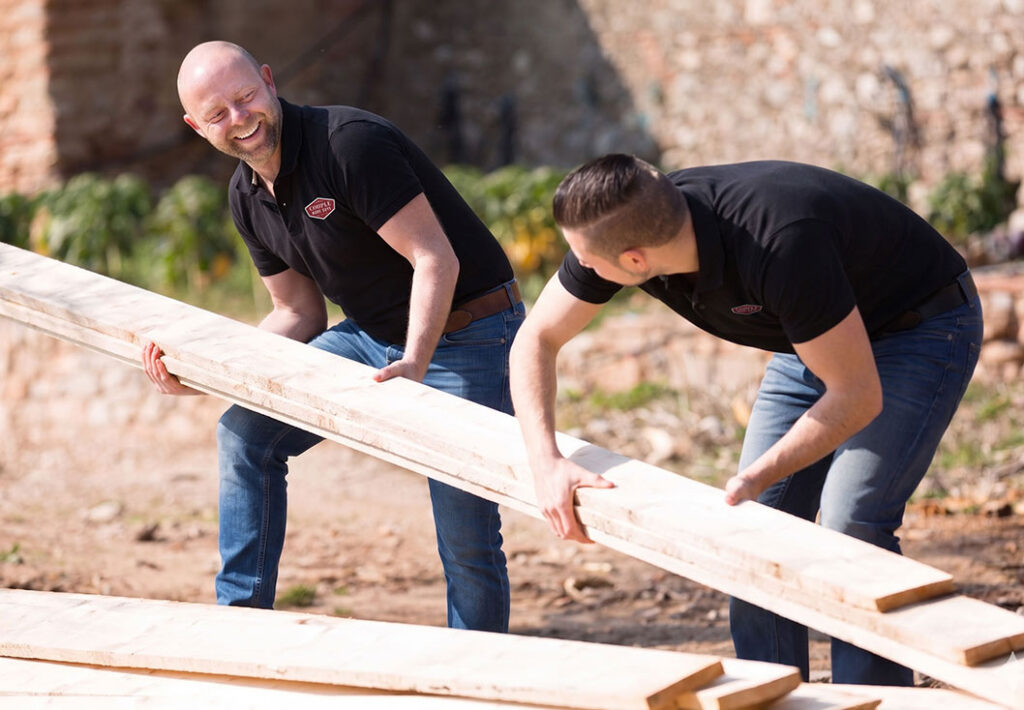 Two men wearing black shirts and jeans are smiling and working together to lift and move long wooden planks outdoors on a sunny day.