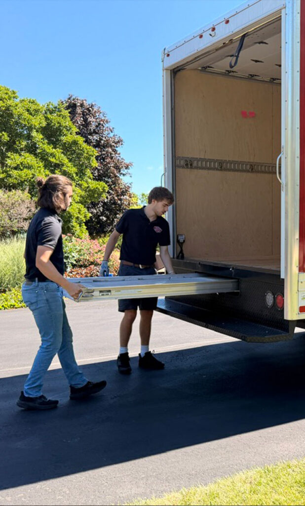 Two people pull out a metal ramp from the back of a large moving truck parked on a sunny driveway, with trees and grass in the background.