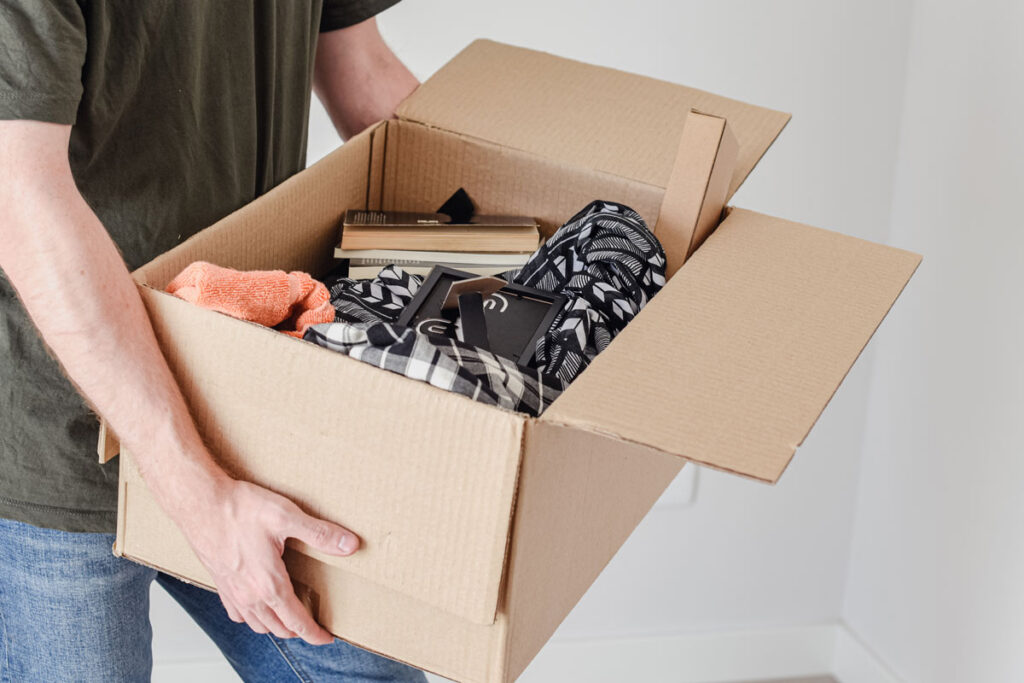 A person holding an open cardboard box filled with folded clothes, books, and various household items, standing indoors against a white wall.
