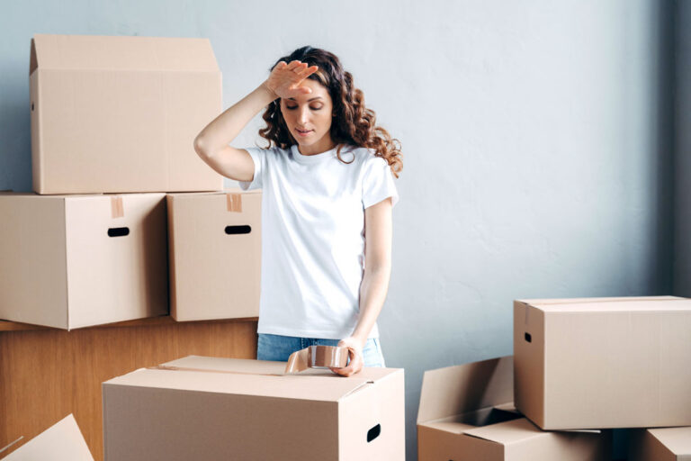 A woman in a white t-shirt stands surrounded by cardboard boxes, wiping her forehead and looking tired, possibly from packing or moving. One hand rests on a large box with packing tape on top.