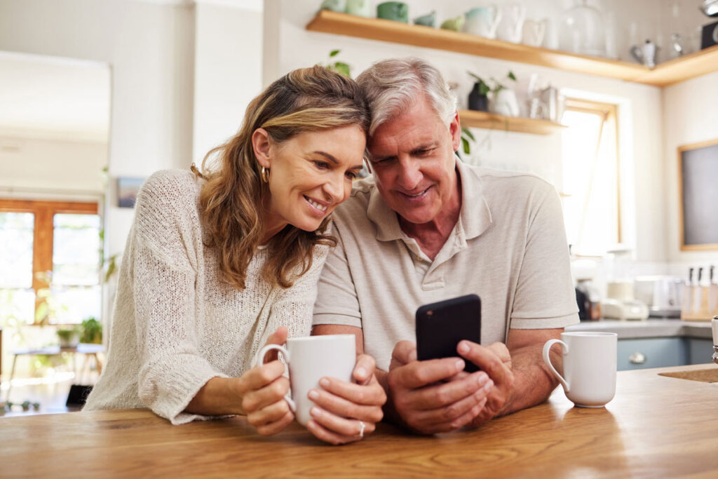 A smiling couple sits at a kitchen counter, holding coffee mugs and looking at a smartphone together, appearing happy and relaxed in a bright, cozy kitchen.