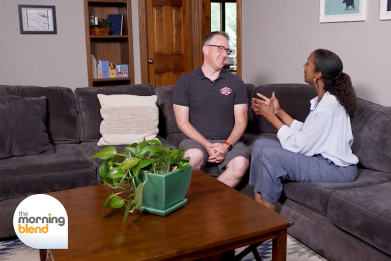 A man and a woman sit on a gray couch, smiling and talking. A green potted plant is on the wooden coffee table. Bookshelves and framed art are in the background. The Morning Blend logo is in the bottom left corner.