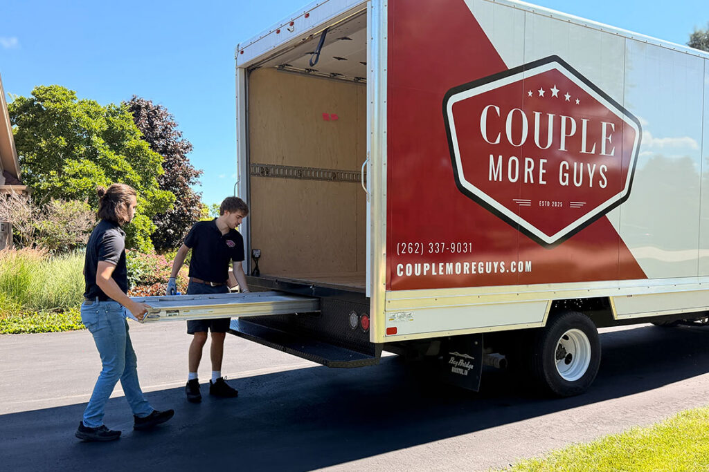 Two people load a large moving truck with Couple More Guys branding on its side. The truck is parked on a driveway with green trees and grass visible in the background.