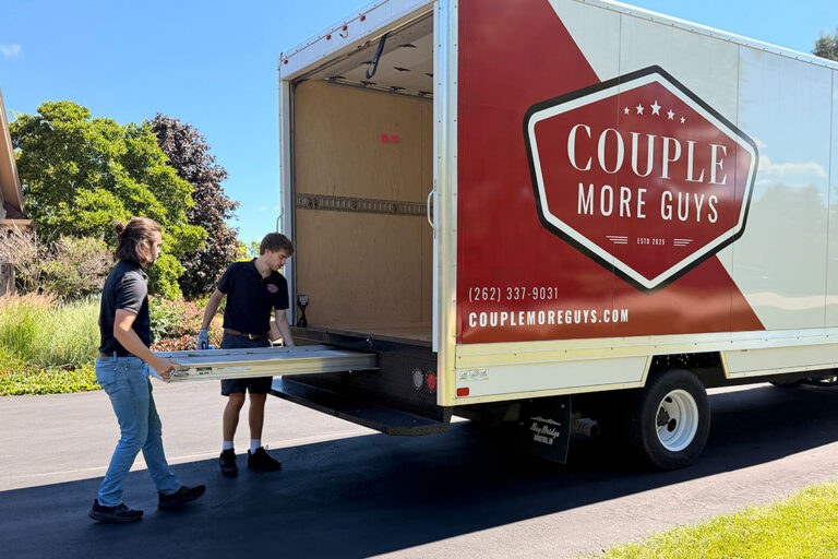 Two people load a large moving truck with Couple More Guys branding on its side. The truck is parked on a driveway with green trees and grass visible in the background.