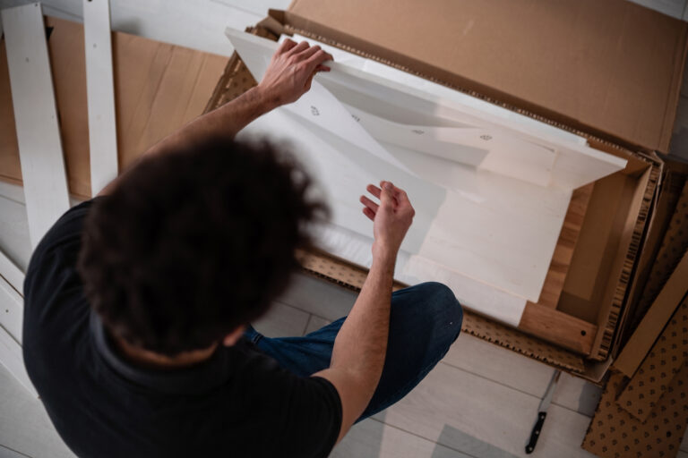 A person sits on the floor, assembling white wooden furniture parts from a box, with screws and a screwdriver nearby. The photo is taken from above, showing their dark hair and casual clothing.
