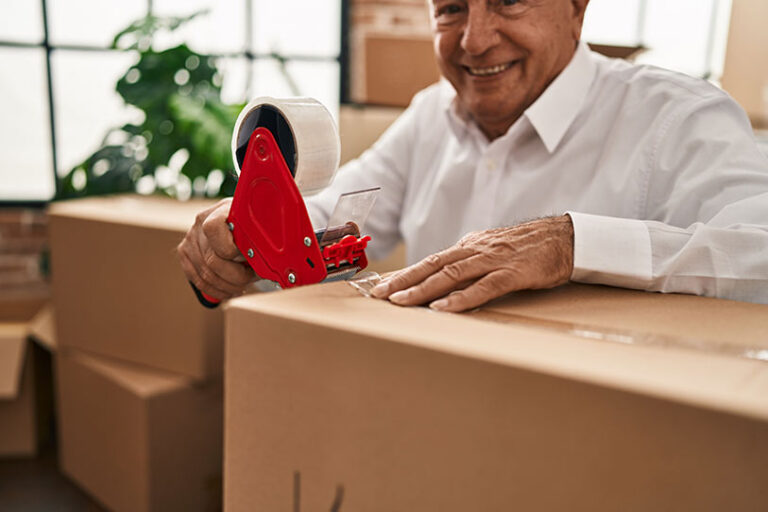 Smiling person in a white shirt seals a cardboard box with clear packing tape using a tape dispenser, surrounded by other boxes, possibly during a move or packing process.